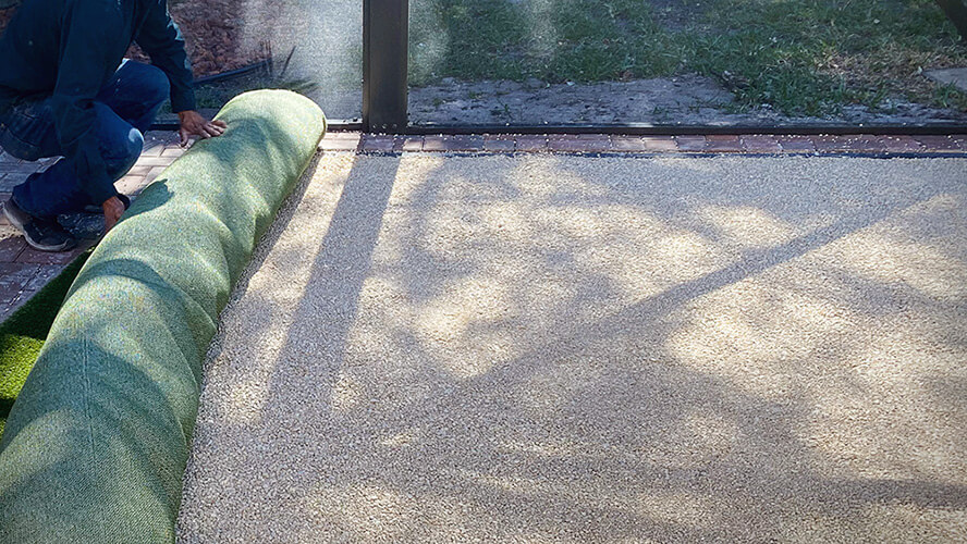 A worker unrolls artificial turf on compressed granite in a backyard space.